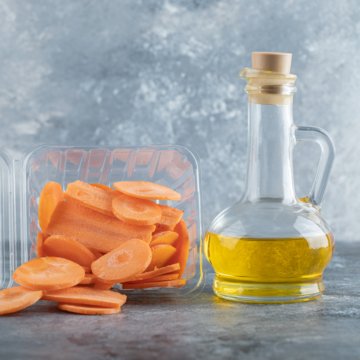 Pile of sliced carrots in plastic container and bottle of oil over grey background
